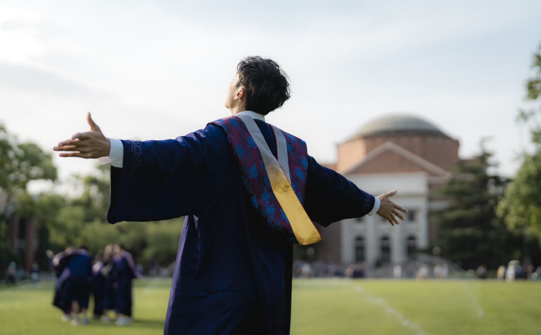 A graduate stands with open arms on a university lawn.